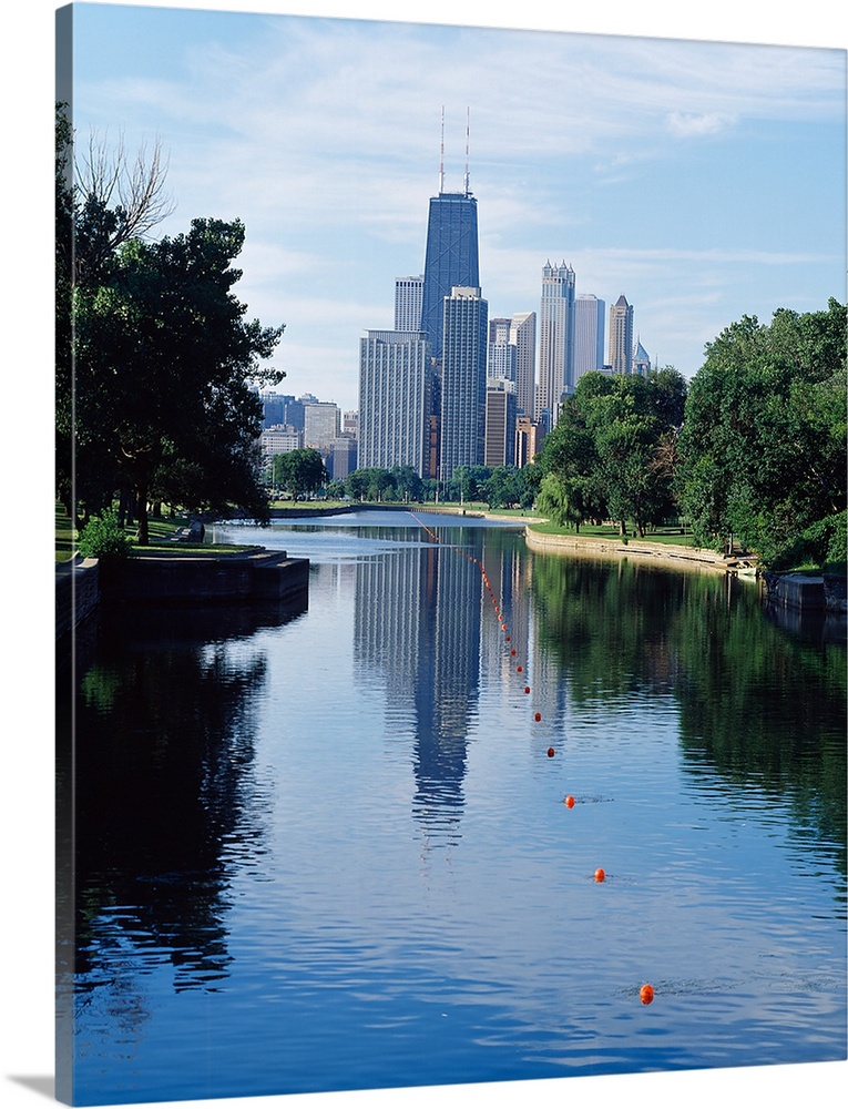 Vertical panoramic photograph of buildings and skyline reflected in pond lined with trees.