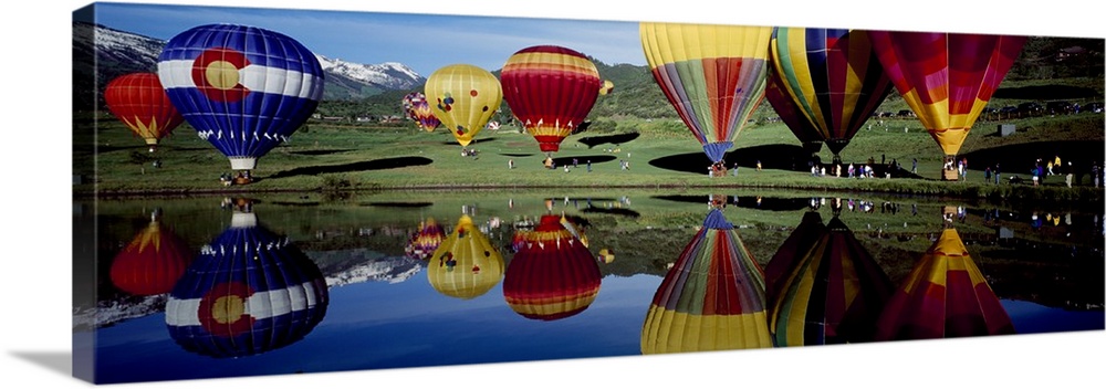 Long photo on canvas of big balloons about to take off in a field that are reflected in the waterfront.