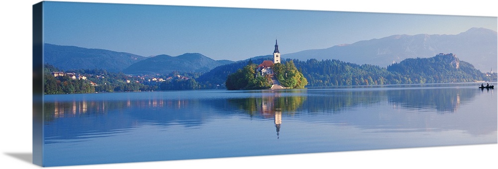 Giant, horizontal photograph of a mountain landscape and buildings reflecting in the waters of Lake Bled, in Slovenia.