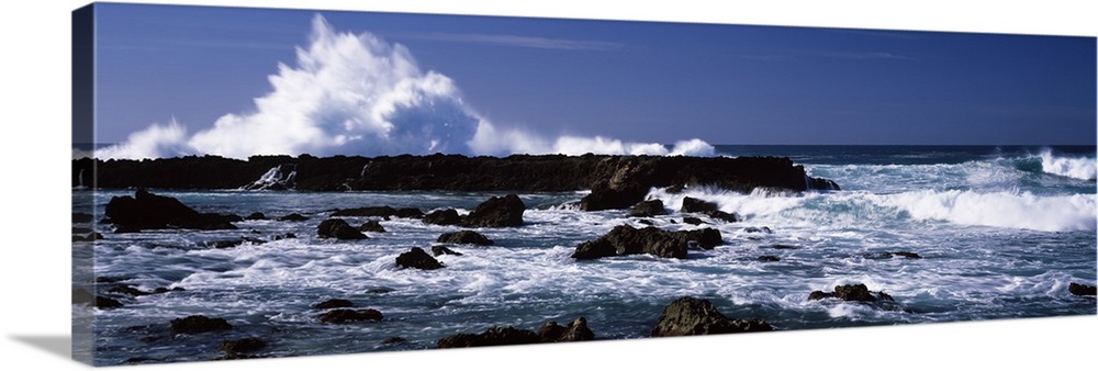 Panoramic photograph of rocks at sea with waves crashing around them under a clear sky.