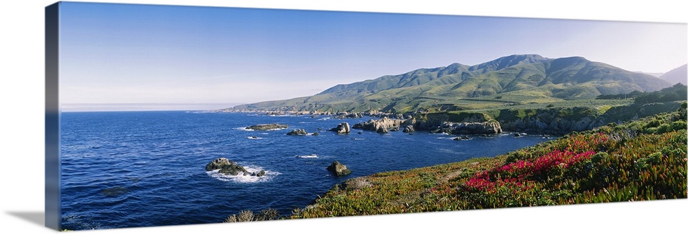 Horizontal, oversized photograph of blue waters with protruding rocks, mountains in the distance, in Carmel, California.