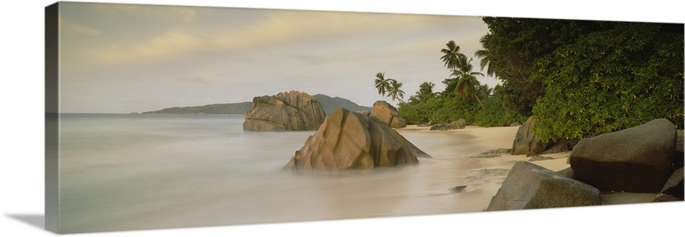 This large panoramic photograph shows a beach with lush green forest behind it and rocks sitting in the water with fog lin...