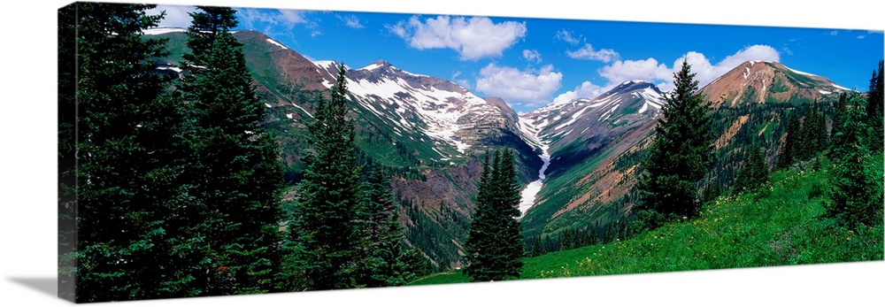 This panoramic photograph taken from the side of a mountain looks across a valley to the snowcapped peaks on the other side.