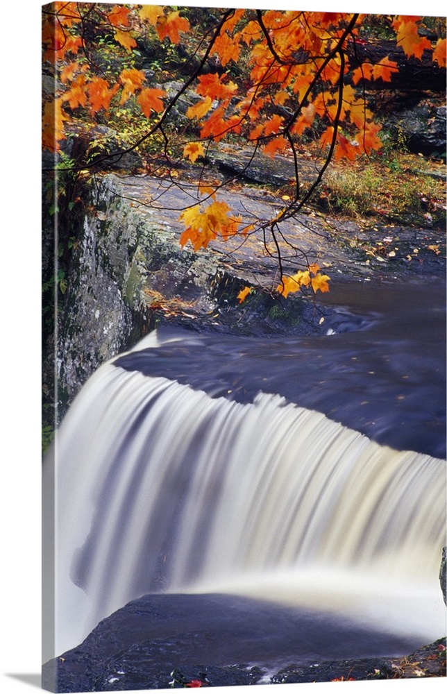 Peaceful scene of a waterfall in a New England forest in the fall, with a rocky cliff and bright orange leaves.