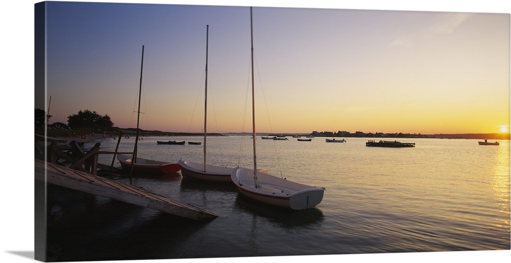 Three sail boats sit docked in the water as the sun sets off in the distance.