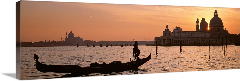 Panoramic photograph of gondola on waterway with cityscape in background at sunset.