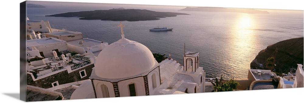 Giant, high angle photograph of the tops of buildings, looking out onto the water in Santorini, Greece as the sun sets ove...