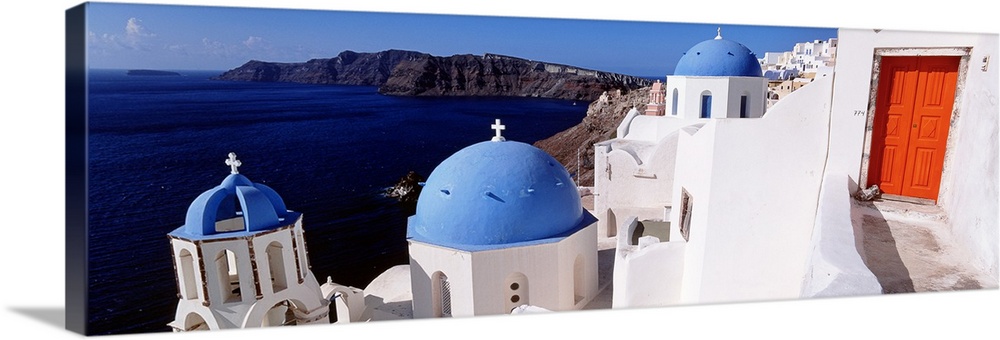 Panoramic photograph of whitewashed buildings with colorful roofs near coastline with mountains in the distance.