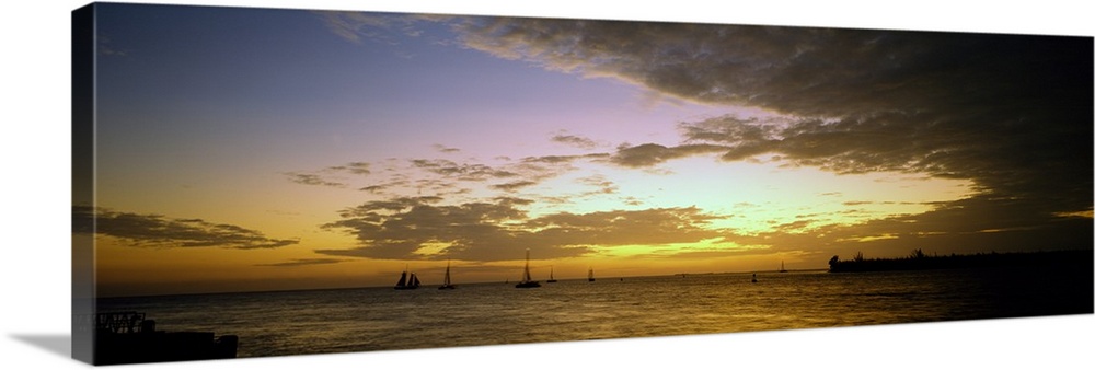 Panoramic photograph of ocean with sailboats in the distance under a cloudy sky at dusk.