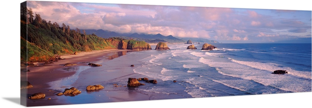 Panoramic photograph of waves slowly crashing against the rocks and sand of Cannon Beach in Oregon.  The mountains in the ...