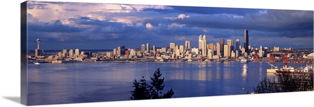 Panoramic, distant photograph over the water of the Seattle skyline, beneath a sky full of large, billowing clouds.  Tree ...