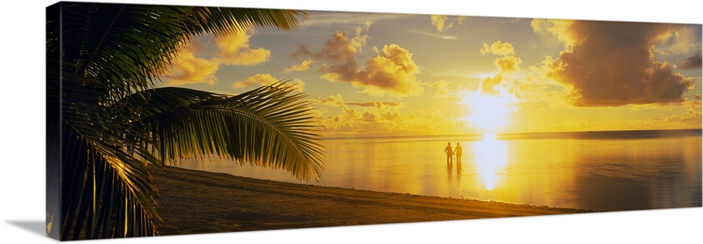 A panoramic photograph of the sun setting over a tropical sea and cloud filled sky with palm trees in the foreground.