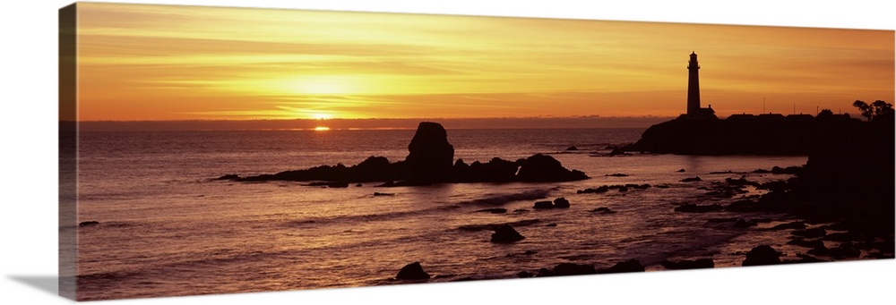 A lighthouse and the cliffs surrounding it are silhouetted by the sunset over the Pacific ocean.
