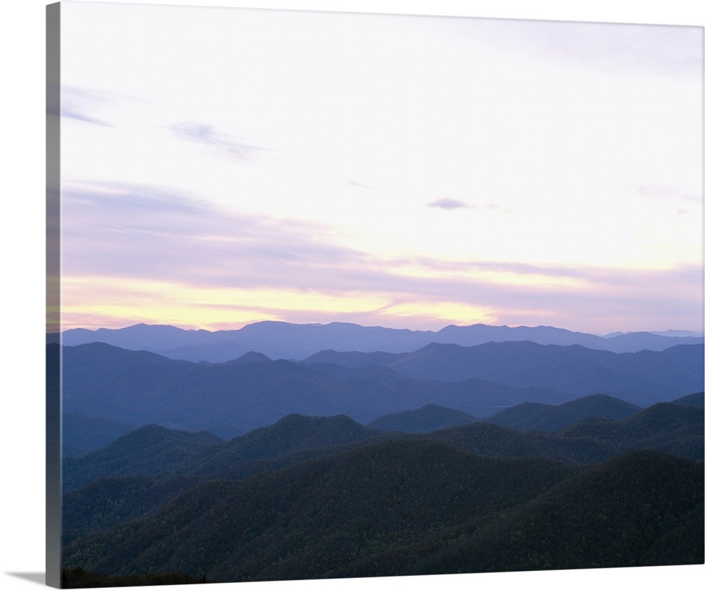 Rows of silhouetted mountains covered in green leaf trees as far as the eye can see as the sun rises in the distance.