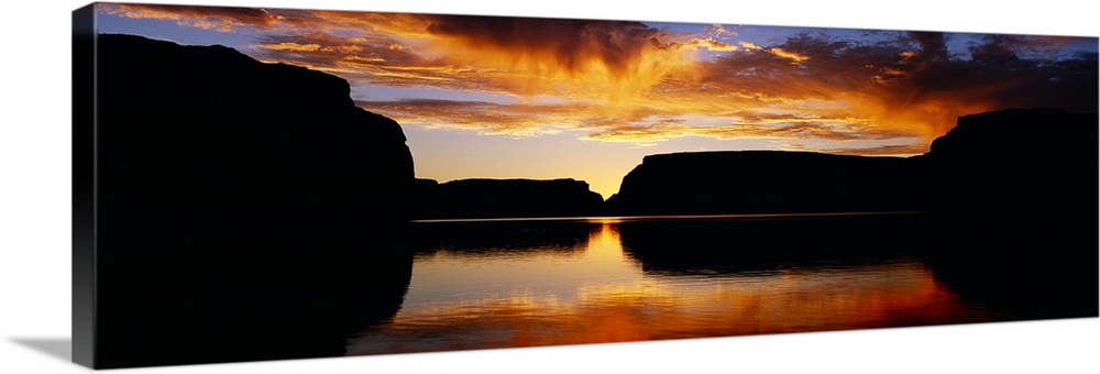 Giant landscape photograph of large, silhouetted rocks surrounding Lake Powell, beneath a vibrant sky at sunset, in Utah.