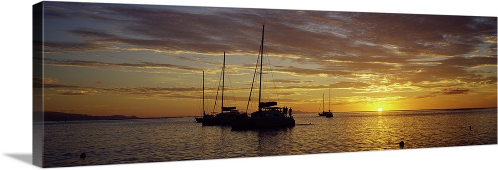 Panoramic photograph on a big wall hanging of several silhouetted sailboats, floating on calm waters as the sun sets on th...
