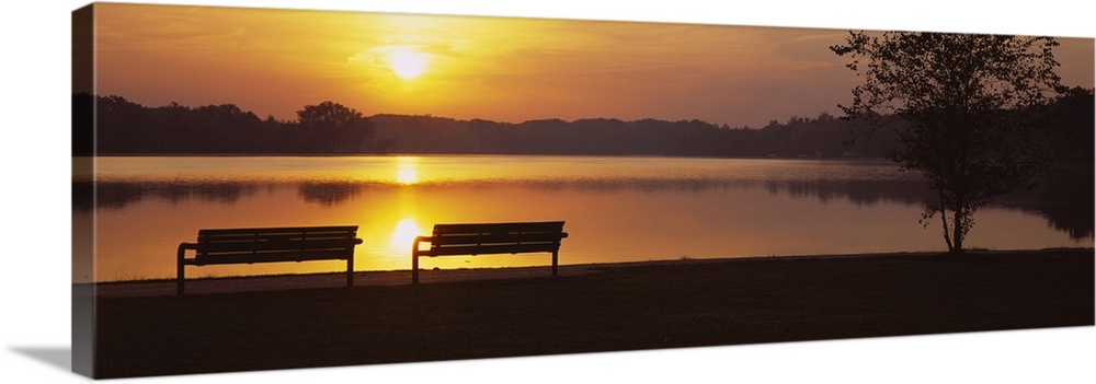 Panoramic photograph of park benches near water's edge at sunset with forest silhouette in the distance.