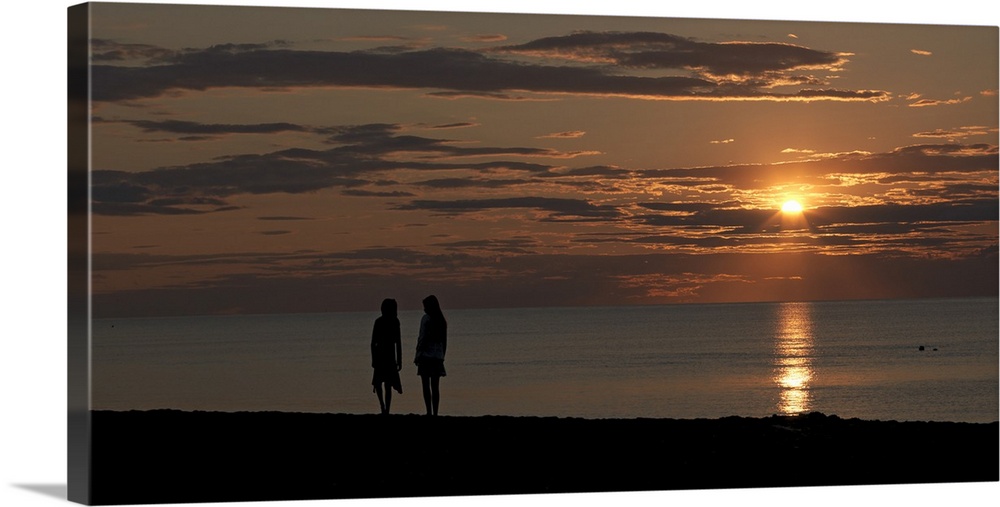 Panoramic photo of the silhouette of two people walking along the ocean at sunset.