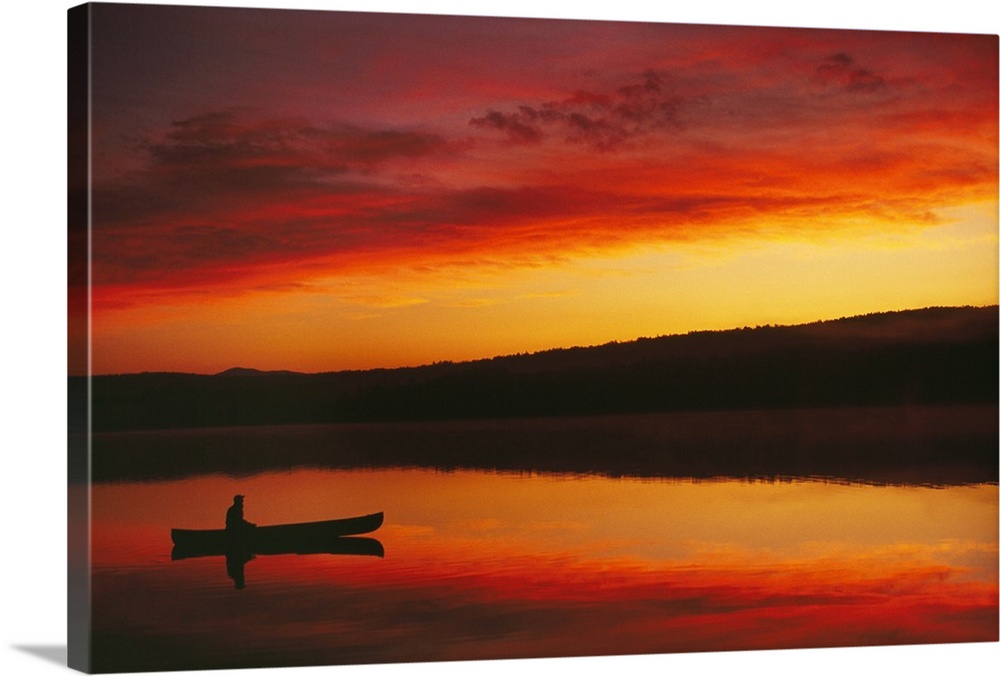 Big, landscape photograph of the silhouette of a person sitting in a boat beneath a vibrant, fiery sunset.