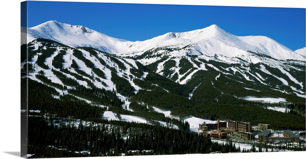 Ski lodges are shown at the bottom of snowy, forested mountain range in this large photograph.