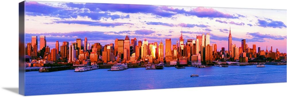 Panoramic photograph of the water in front of the New York City skyline, beneath a vibrant, partly cloudy sky at sunset.