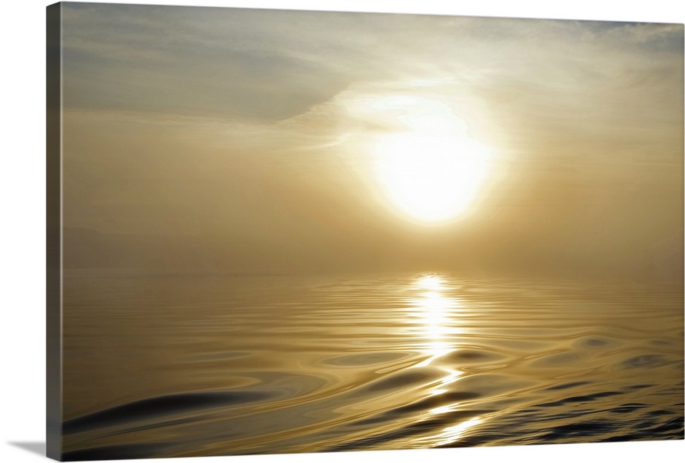 Large landscape photograph of a rising sun covered by a thin haze in the sky, above the rippling, smooth waters of Waterfo...
