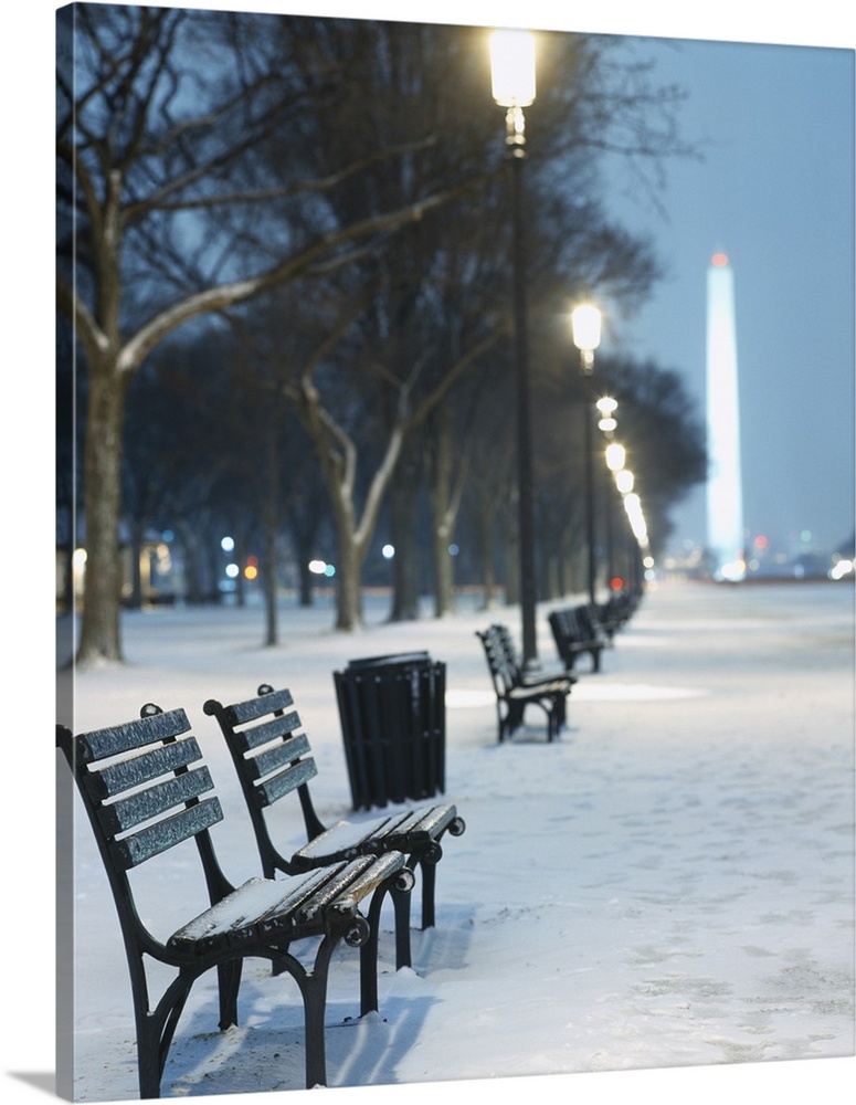 Snow covers the National Mall and rows of out of focus benches and street lights in this vertical landscape photograph.