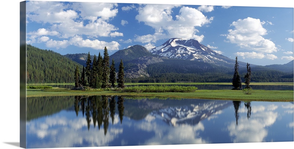 Panoramic photograph of lake front meadow with trees and shrubbery.  A forest is in the background with snow covered mount...
