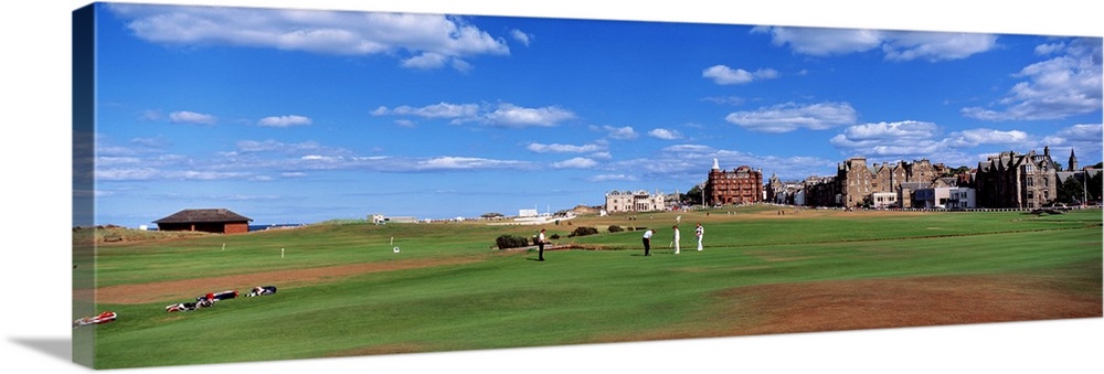 A group of people tee off on a golf course in the UK. Buildings on the course line the right side of the panoramic piece.