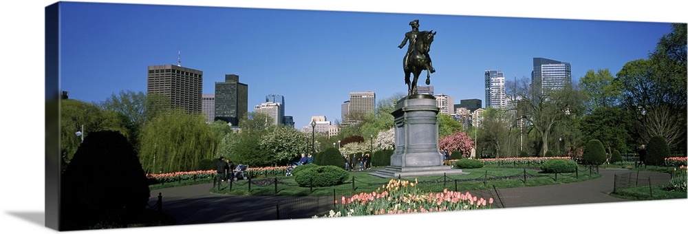 A large statue that stands in the middle of a garden is photographed in panoramic view with tall buildings visible in the ...