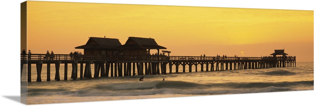 A silhouetted wooden wharf with small buildings stretches out into the ocean at sunset. A couple surfers float in the wate...