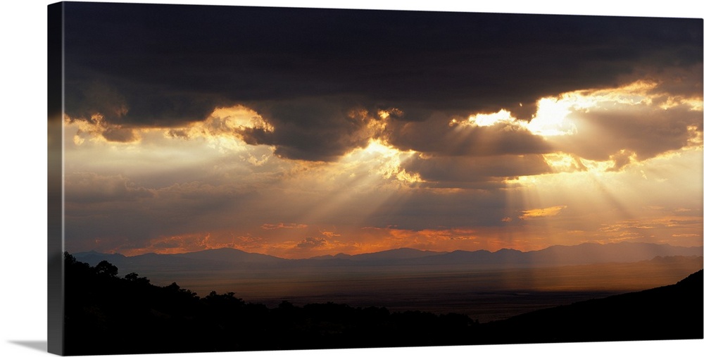 Horizontal, large photograph of sunlight beaming through a sky full of huge, dark clouds, over the Chiricahua National Mon...