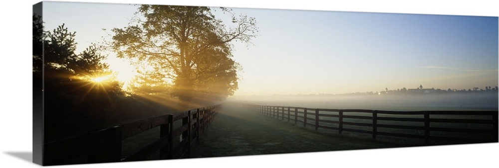 Beams of sunshine reaching across two wooden fences of corrals on a hazy, misty morning in the countryside.