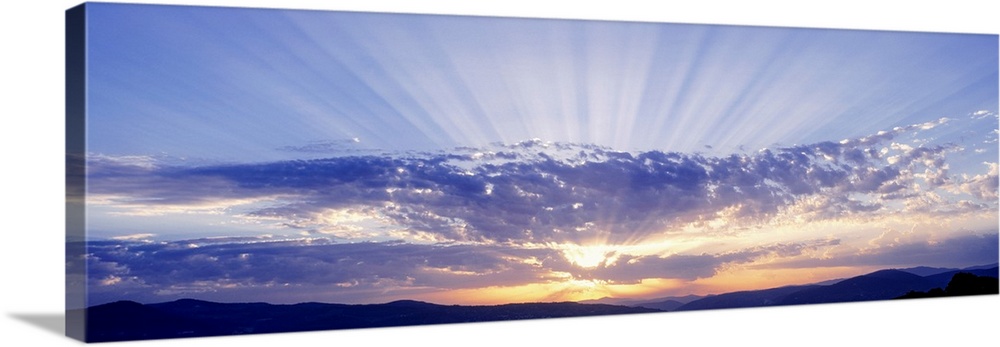 This panoramic wall art captures as a photograph radiant sun beams radiate from behind clouds over a hilly landscape.