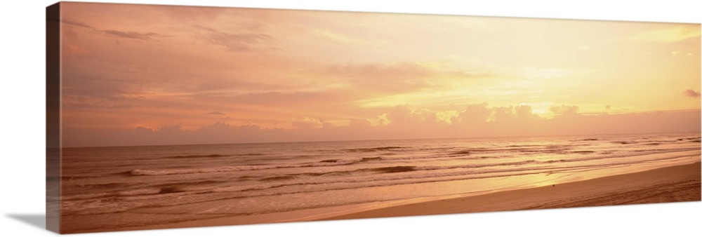 Panoramic photograph of the sun rising above the clouds as small waves break on the sandy shore.