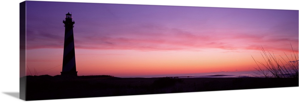 A panoramic canvas with a pastel colored sky with a light house towering over the sandy beach on the left side of this pho...