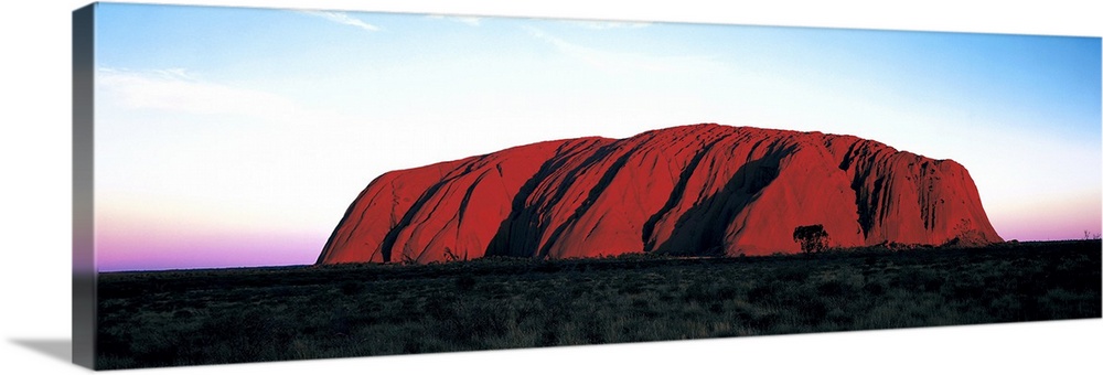 The Ayers Rock is photographed in wide angle view during a sunset which lines the horizon with warm colors.