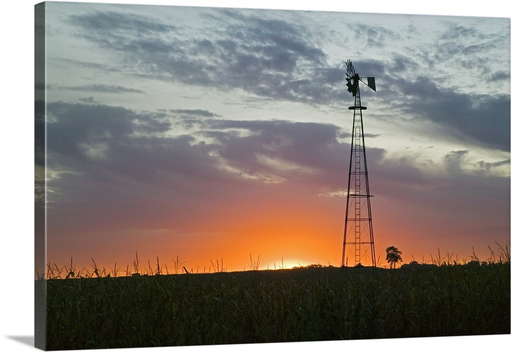 A tall windmill rises above a crop field on a farm with just the edge of the setting sun peeking over the horizon.
