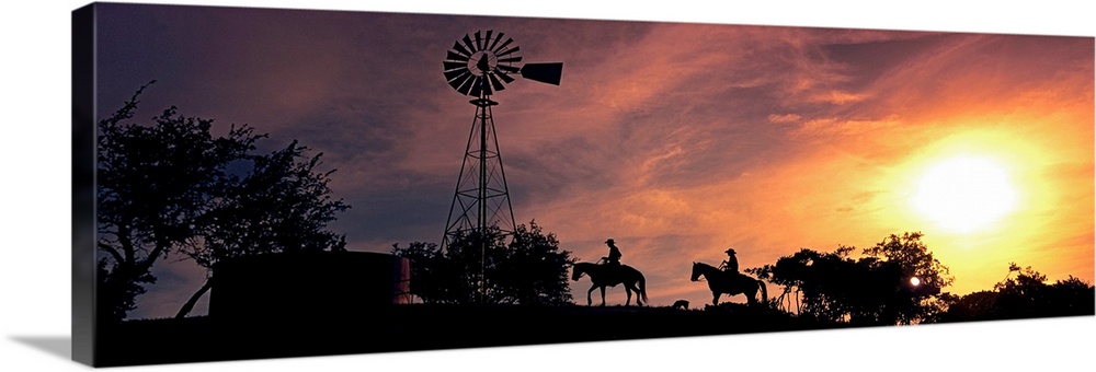 This panoramic photograph depicts an evening moment on ranch hill side as horseback riders pass by a windmill trees.