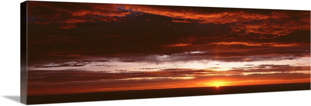 Panoramic photograph of a sunset just hitting the horizon and lighting up a cloud filled sky with warm tones.