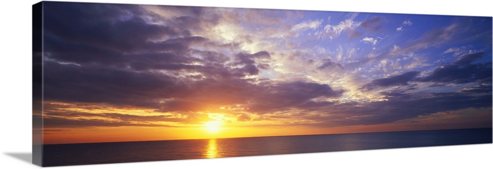 This large panoramic picture was taken of a sunset about to hit the horizon over the ocean with clouds floating in the sky.