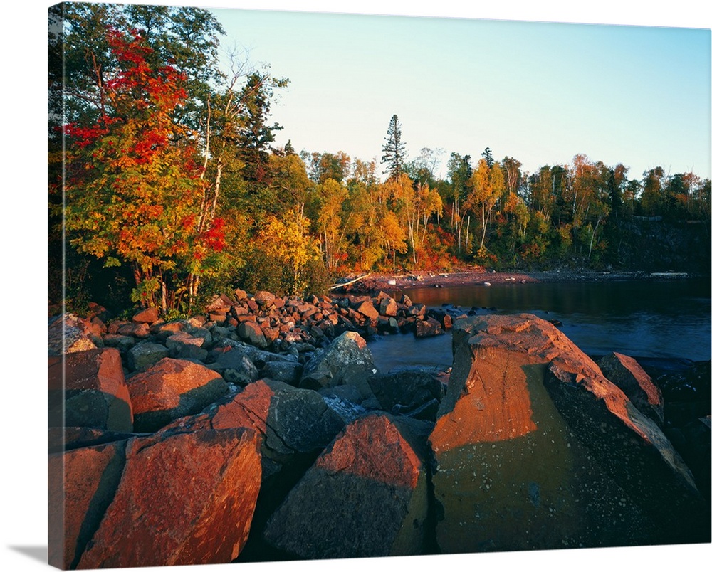 The sun illuminates rust colored rocks and autumn trees at a rocky lake side beach in this landscape photograph.