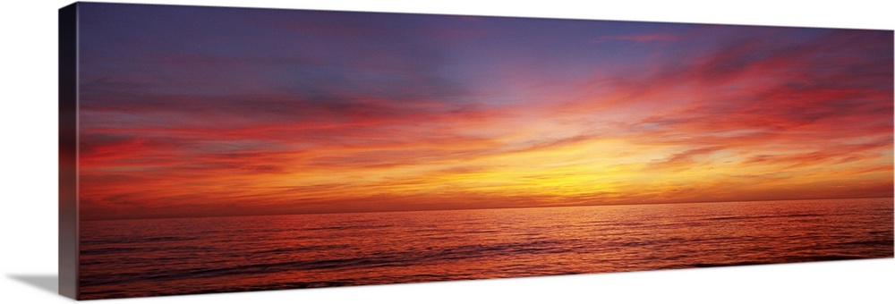 A panoramic photograph of the sun descending behind clouds over the ocean.
