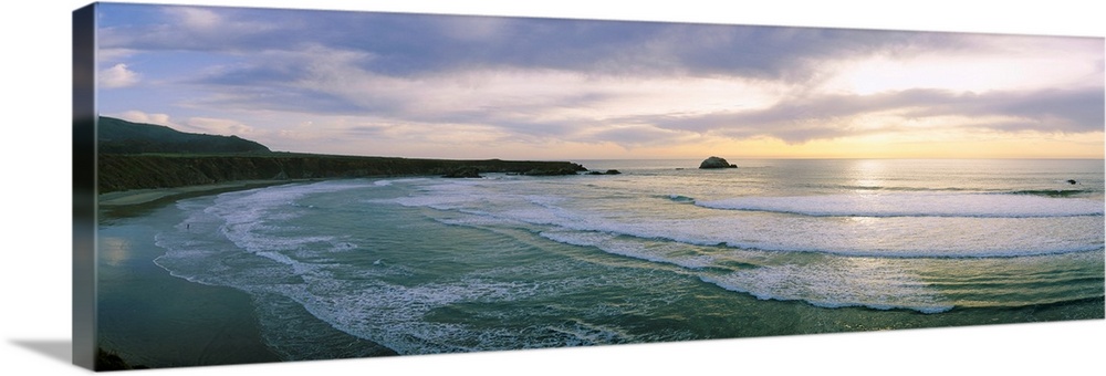 This panoramic wall art is a wide angle photograph of waves washing on to a beach taken from a cliff above the shore.