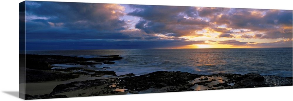 A panoramic photograph of the sun setting beyond the horizon as viewed from a rocky tropical beach.
