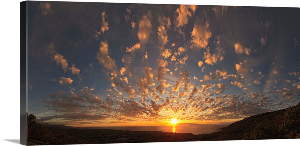 Panoramic photograph taken from the coast of Kona, Hawaii of the sun setting over the Pacific Ocean, beneath a sky of smal...