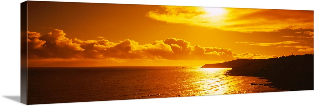 This photograph is a panoramic seascape of the sun reflecting off the shallow beach water as it sinks lower into the horizon.