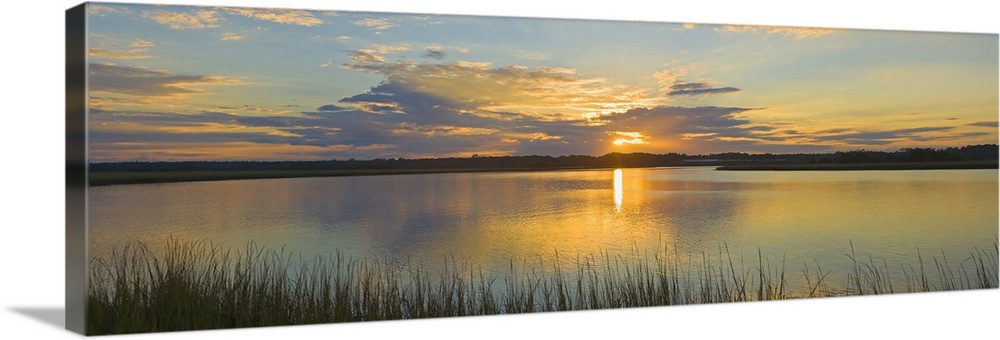 Tall grass lines the bottom of this picture with a wide angle view of a body of water as the sun begins to set over it.