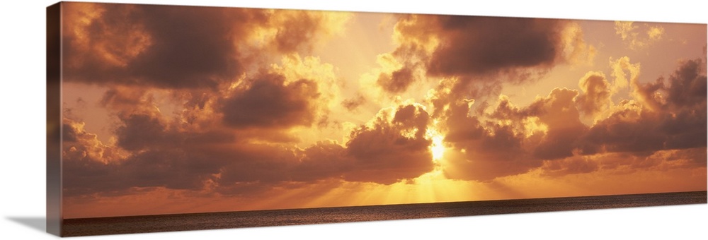 Large panoramic photograph taken of a sunset behind the clouds with its rays bursting through and a vast ocean beneath it.