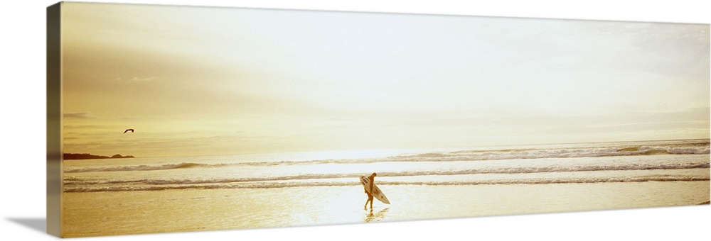 Wide angle photograph of a lone surfer carrying a surf board along Ocean Beach as the tide rolls in at sunset.